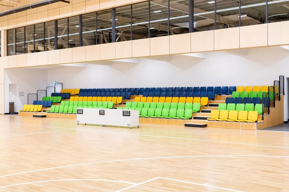 "Indoor fixed bleachers with wooden-look flooring, colorful seats (yellow, green, blue), and a metal structure base featuring fully enclosed treads and backrests."